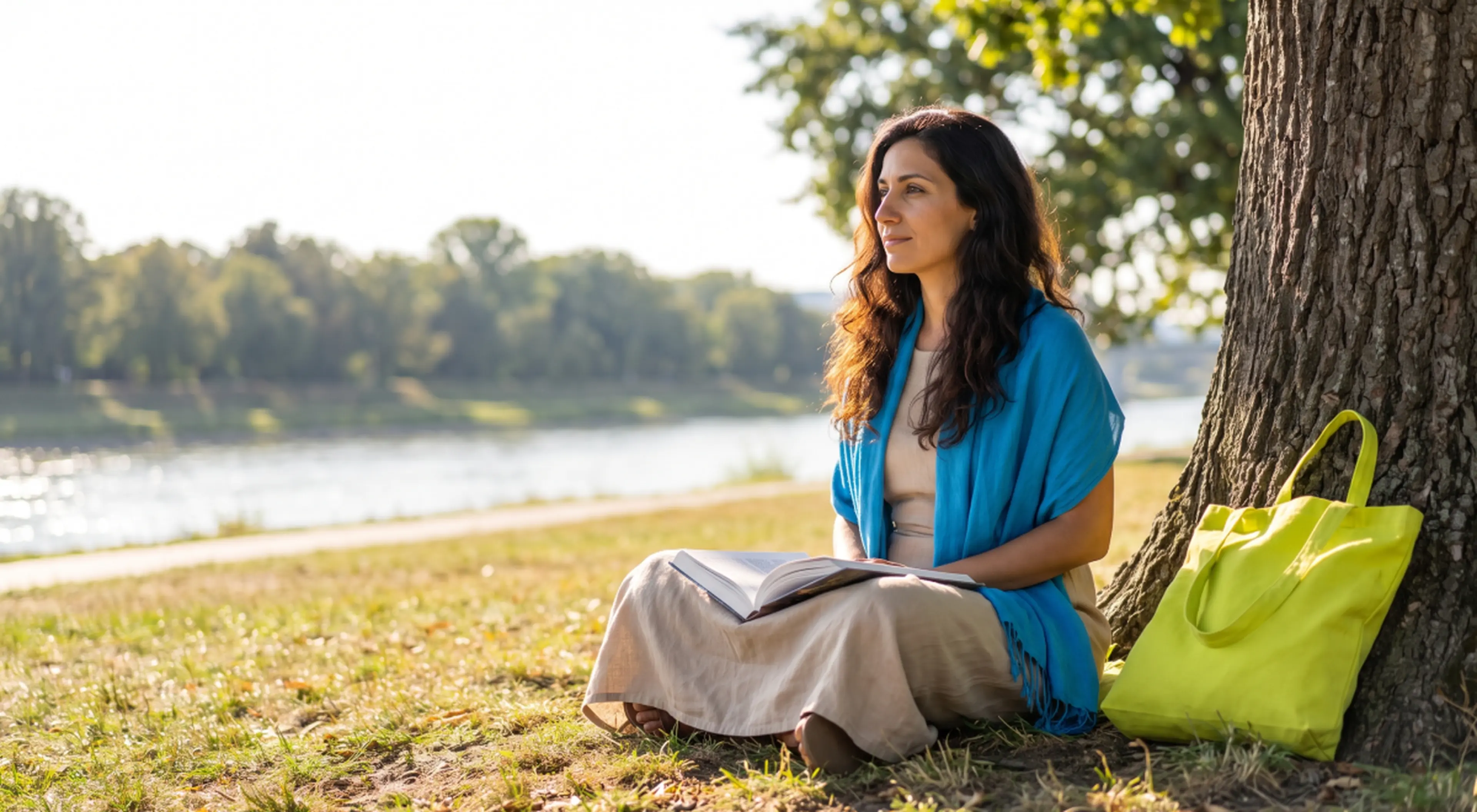 Woman in a blue scarf sitting under a tree by a river with an open book and green tote bag, gazing peacefully as if choosing to disappear for 3 months in nature