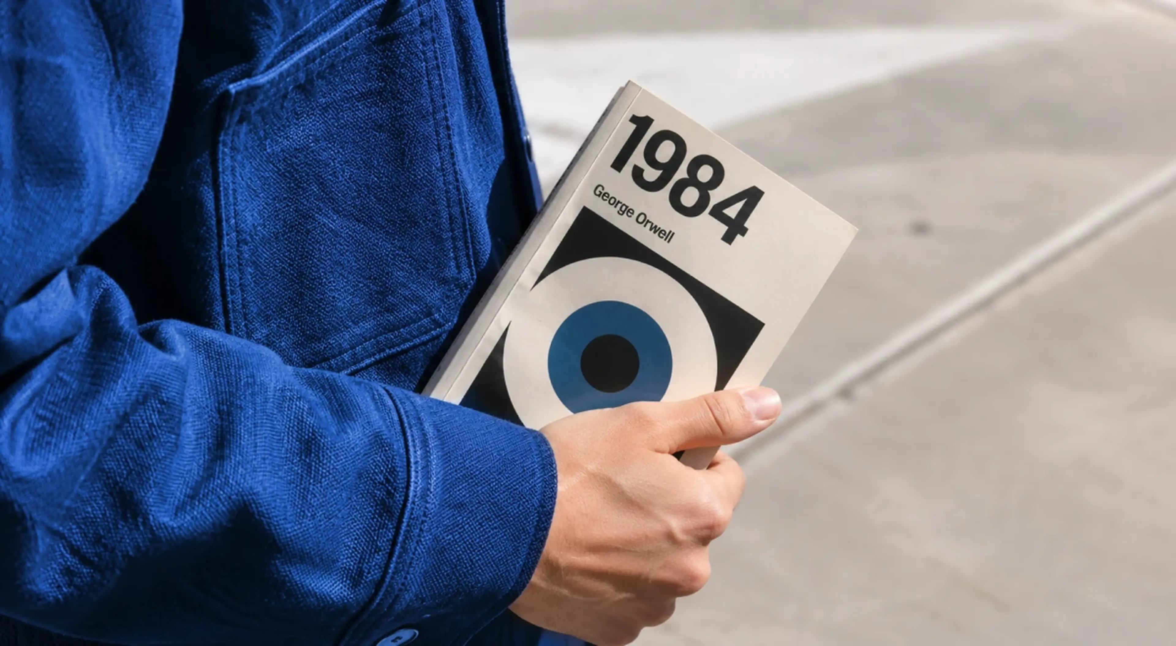Person in a blue jacket holding a copy of 1984 by George Orwell outdoors on a concrete surface, featured among must-read books before you die