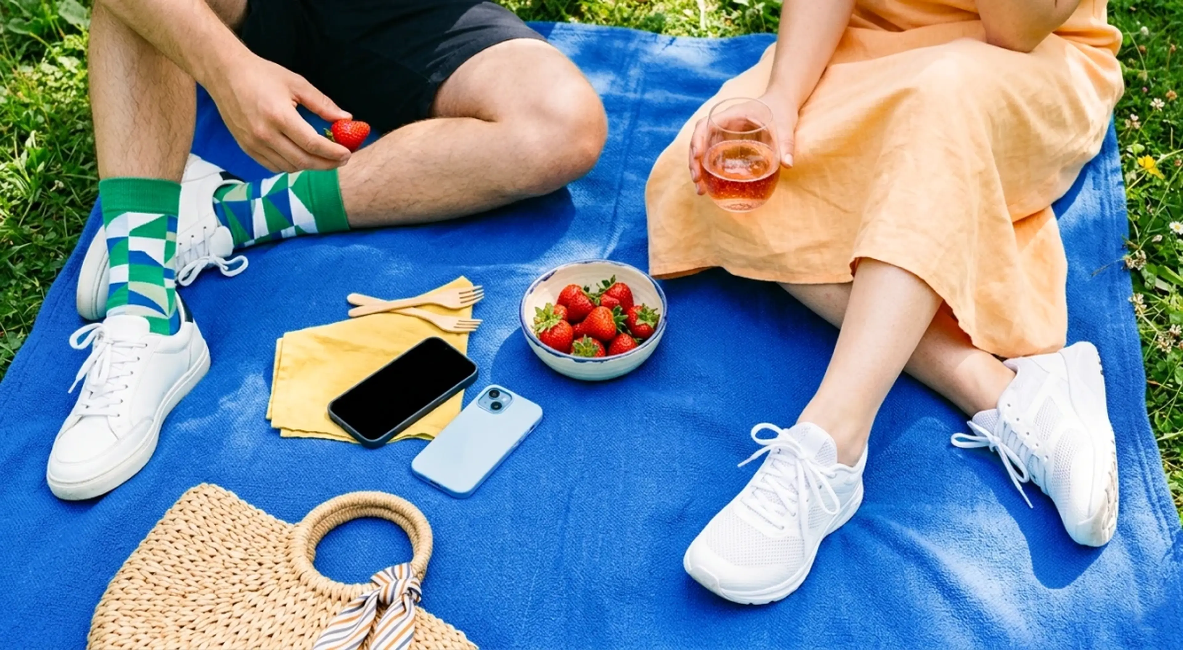 Two people sitting on a blue picnic blanket outdoors, sharing strawberries and rosé wine, with a smartphone, wicker bag, and yellow napkins on green grass background