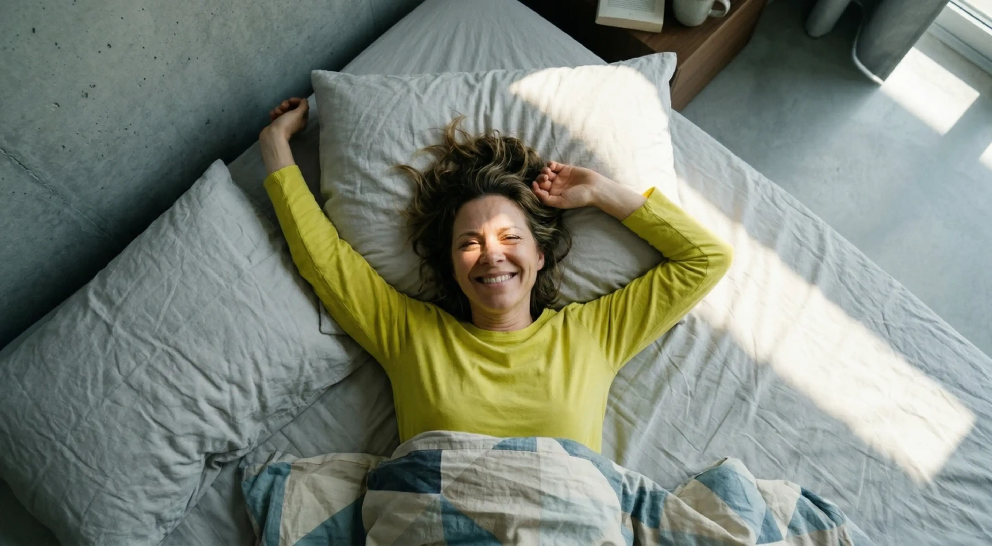 Woman in a yellow top lying on a bed with arms stretched overhead, smiling up at the camera in a bright room, embracing a positive morning routine for well-being