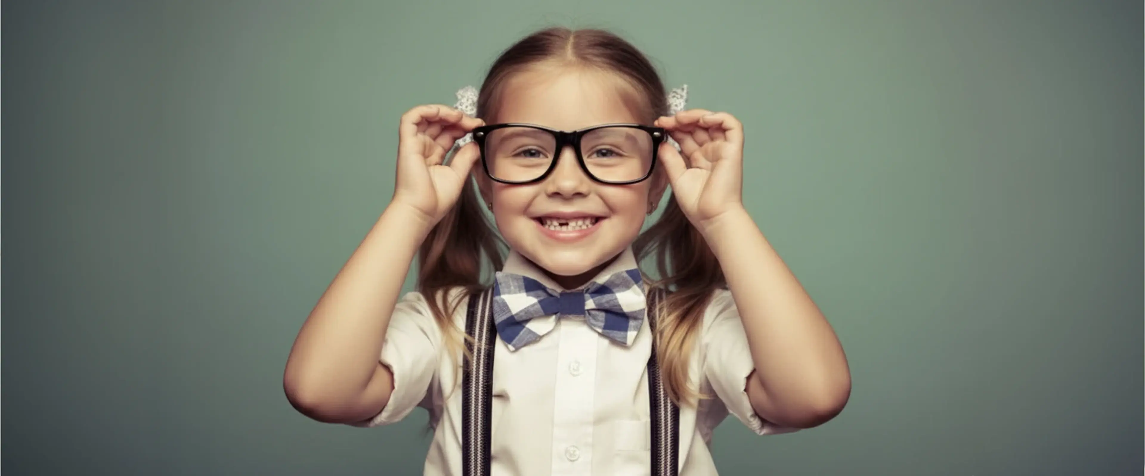 A smiling young girl adjusting oversized black glasses, wearing a bow tie and suspenders, against a teal background, symbolizing intelligence and the limits of IQ measurement