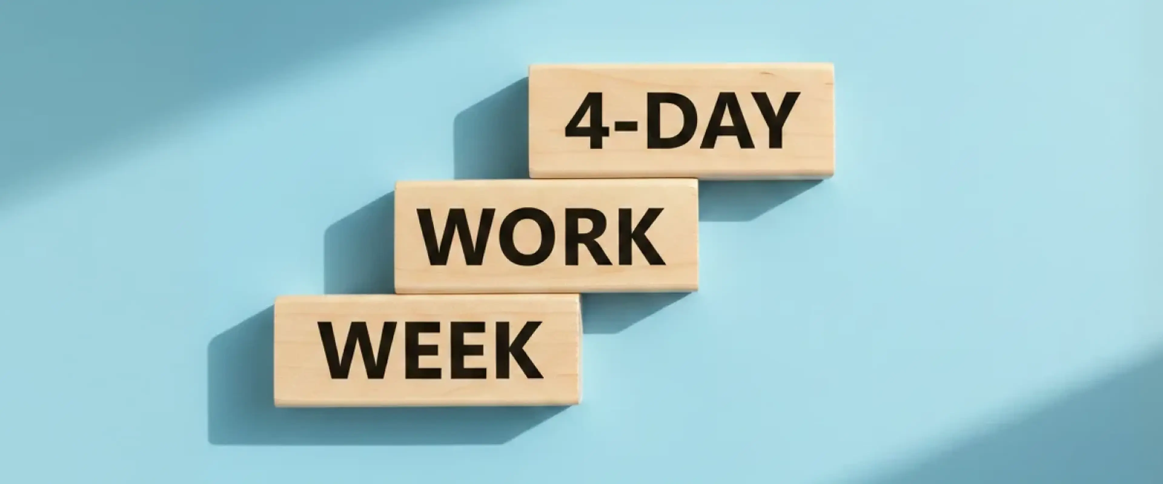 Three wooden blocks arranged in stair-step formation on a light blue background, each labeled to spell out _4-day work week,_ symbolizing the shift to a shorter, more productive work schedule
