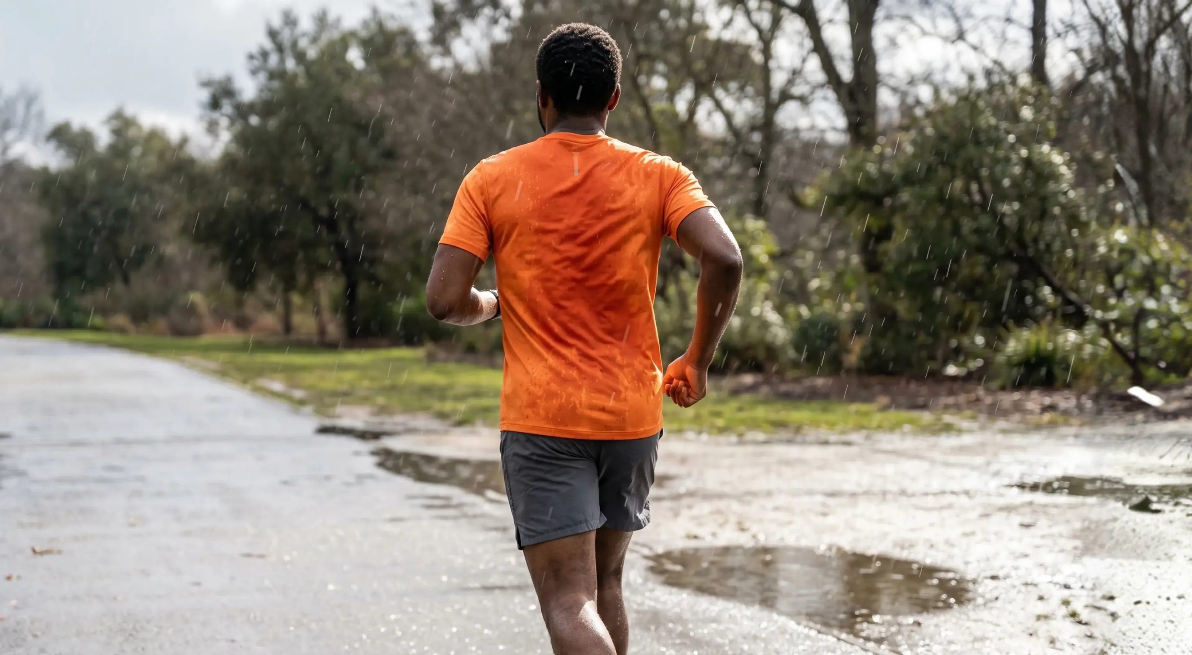Man in orange t-shirt and gray shorts running outdoors in the rain on a wet road surrounded by trees and puddles, demonstrating self-discipline and consistent workout habits