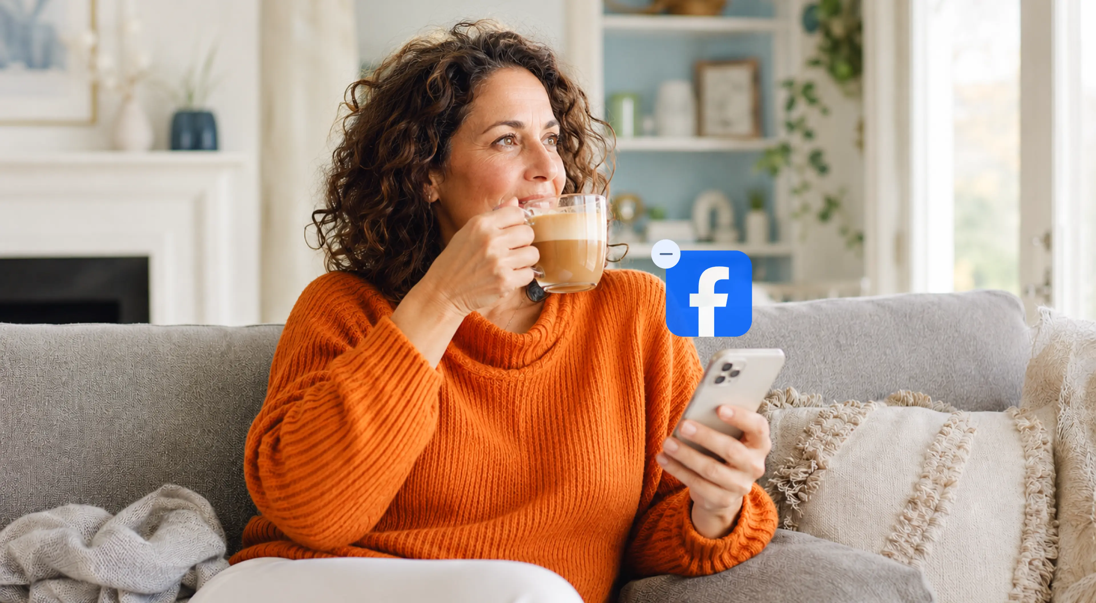 Smiling woman in an orange sweater drinking coffee on a couch while holding a smartphone with the Facebook app icon, in a cozy bright living room