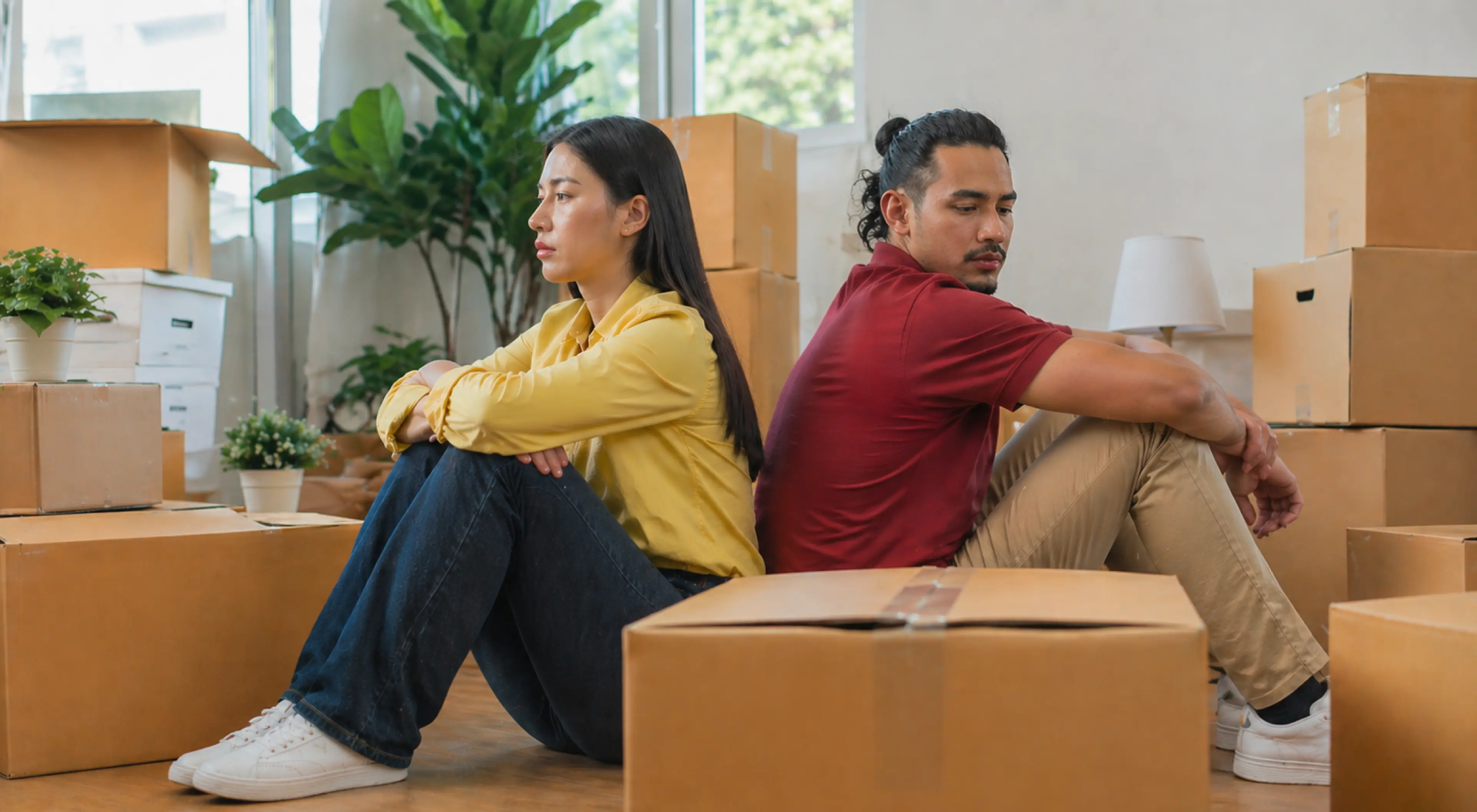 Couple sitting back-to-back on the floor surrounded by cardboard moving boxes, looking away from each other, depicting stages of a breakup in a relationship