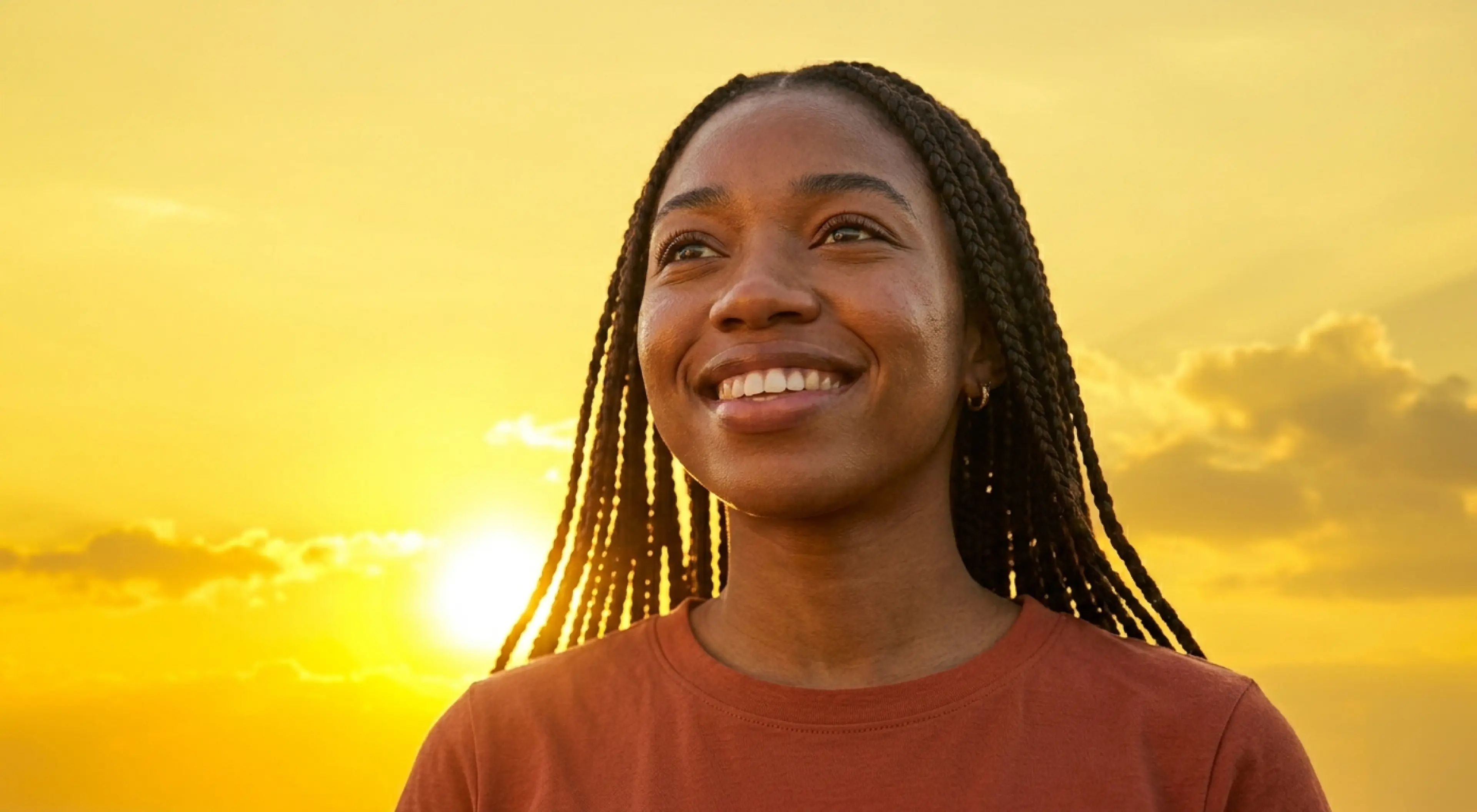 Smiling young woman with braids looking up at a golden sunset sky, radiating positivity and motivation mindset