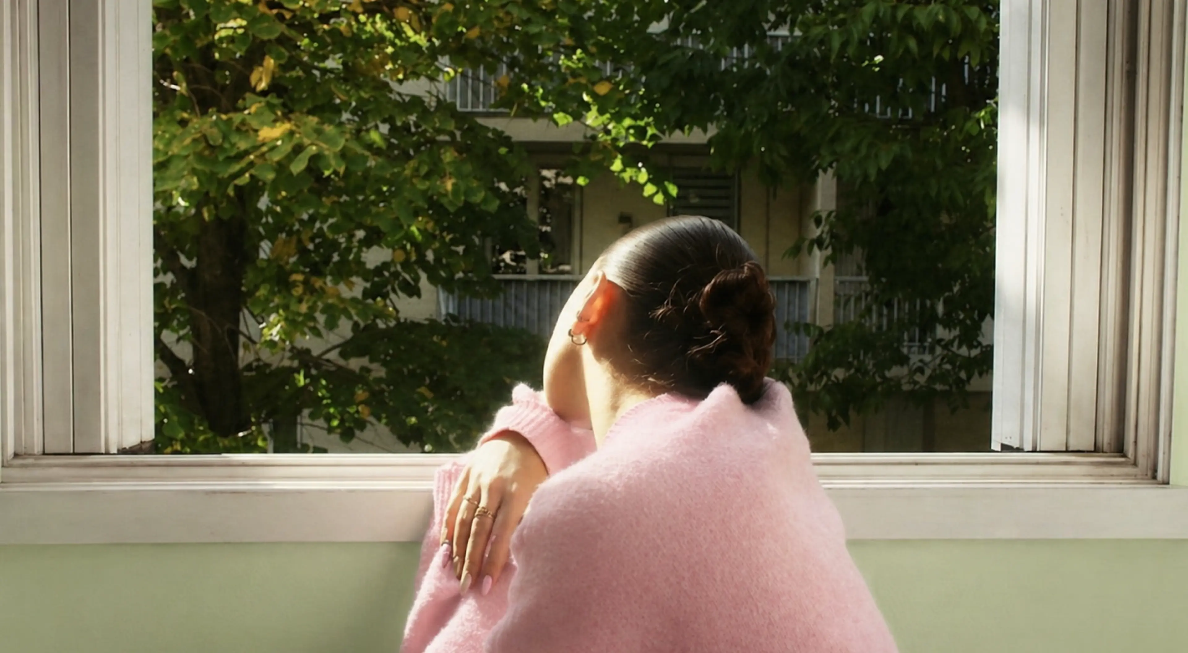 Woman in a pink fuzzy sweater gazing out a sunlit window at green trees, evoking quiet reflection on attachment and the emotional weight of a break up