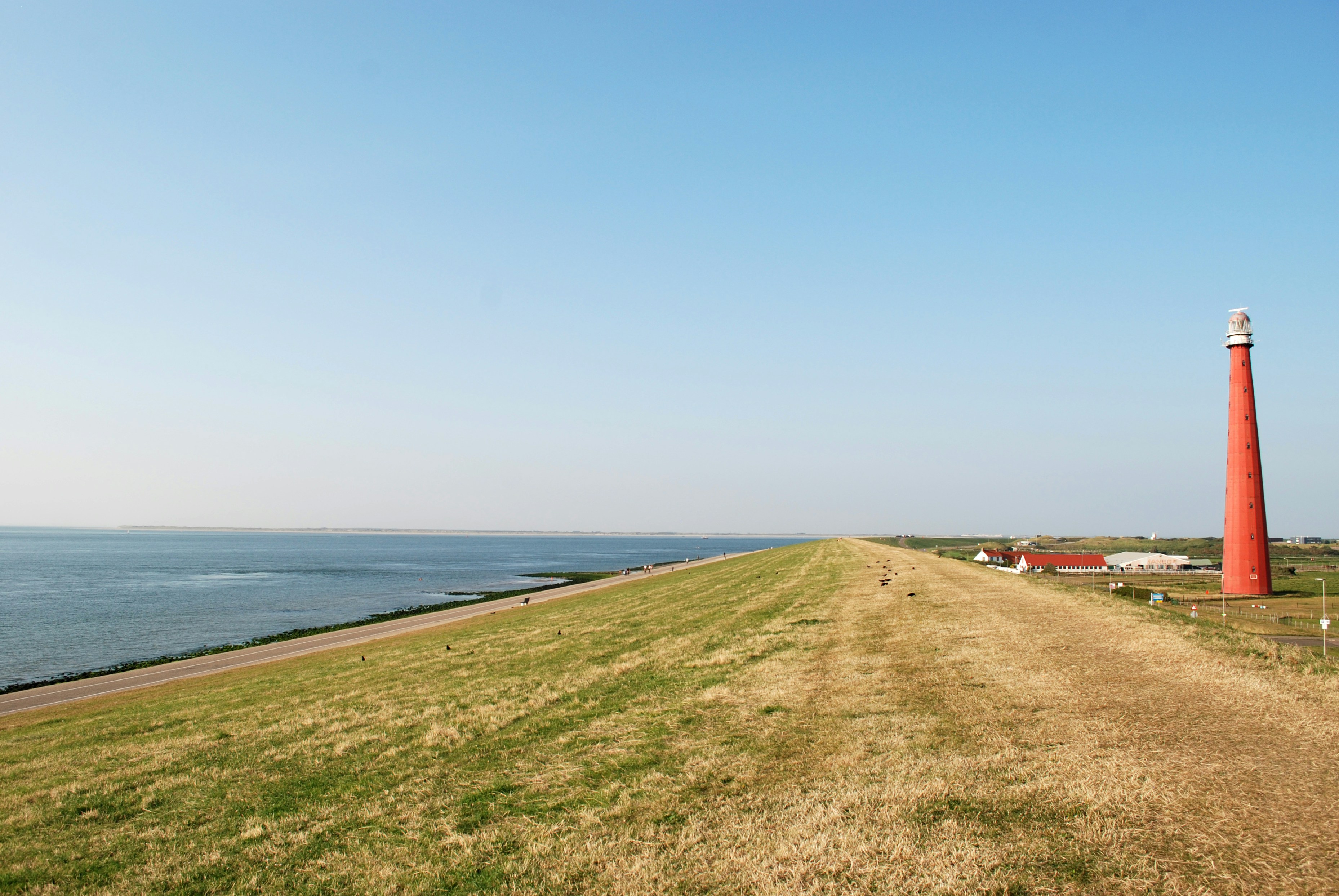 Een rode vuurtoren op een met gras begroeide heuvel, met een weg langs een kalme zee onder een helderblauwe lucht. Op de achtergrond zijn witte huizen te zien.