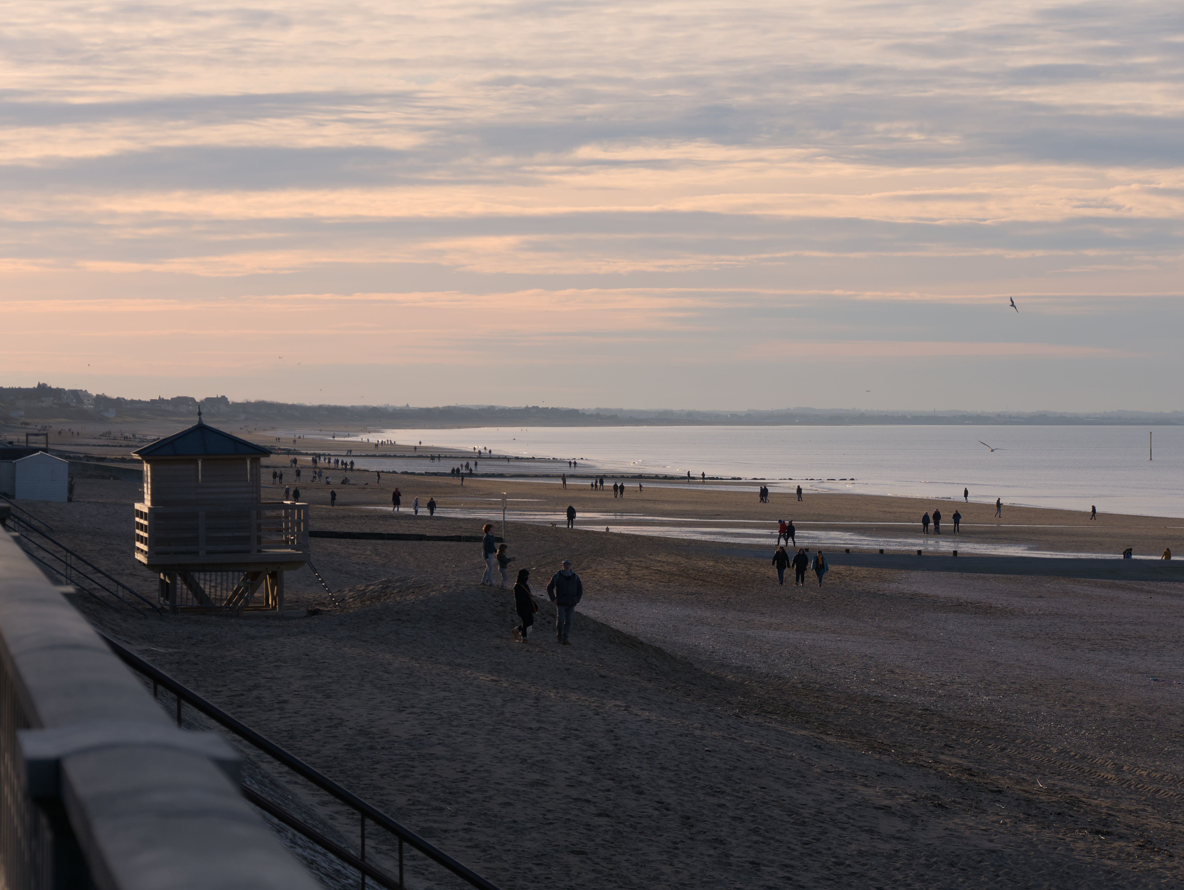 Mensen die bij zonsondergang over een zandstrand wandelen, met een houten uitkijkpost voor strandwachten op de voorgrond en een kalme zee onder een bewolkte hemel op de achtergrond.