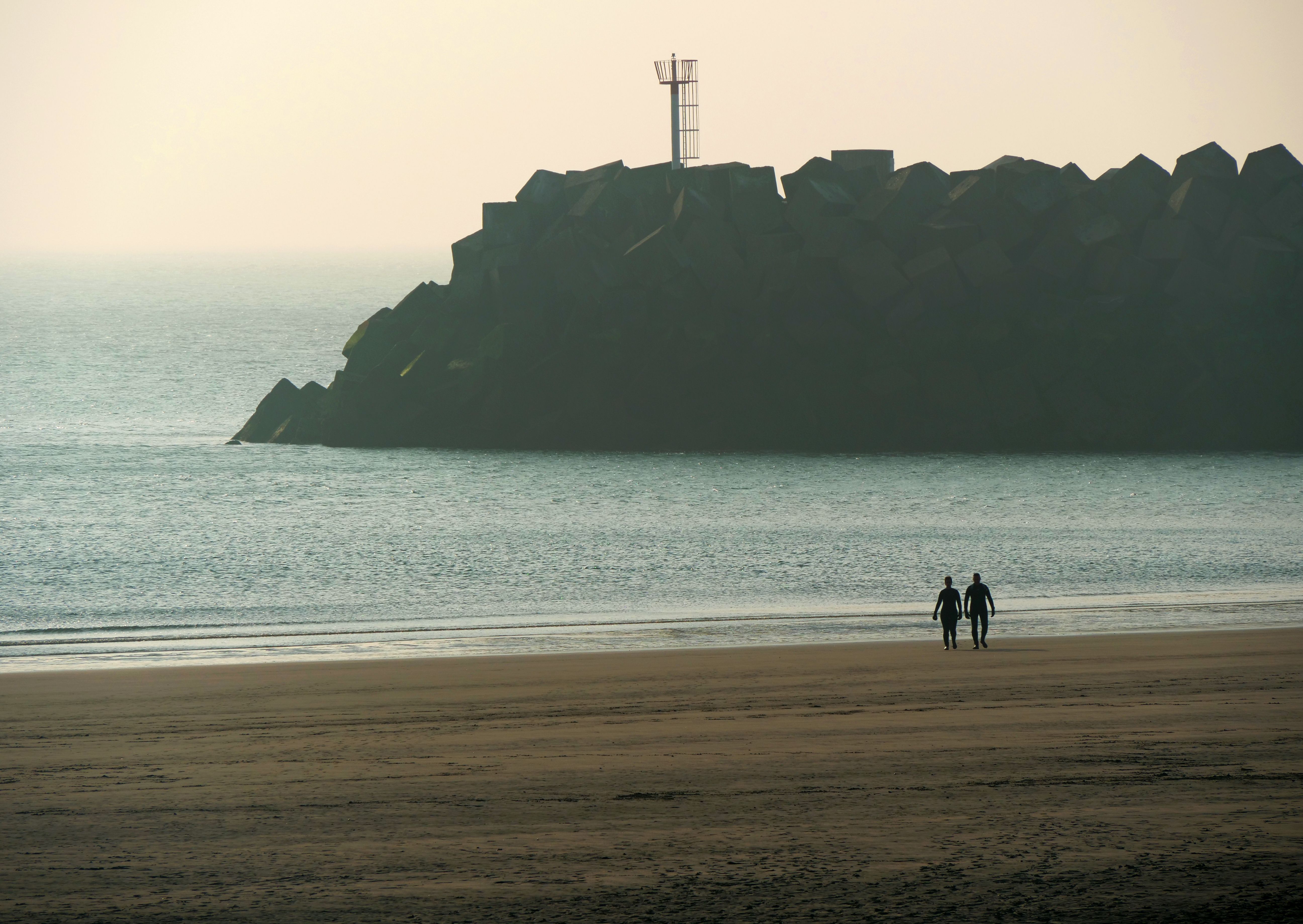 Twee mensen lopen over een zandstrand richting de oceaan, met een rotsachtige golfbreker en een nevelige horizon op de achtergrond.