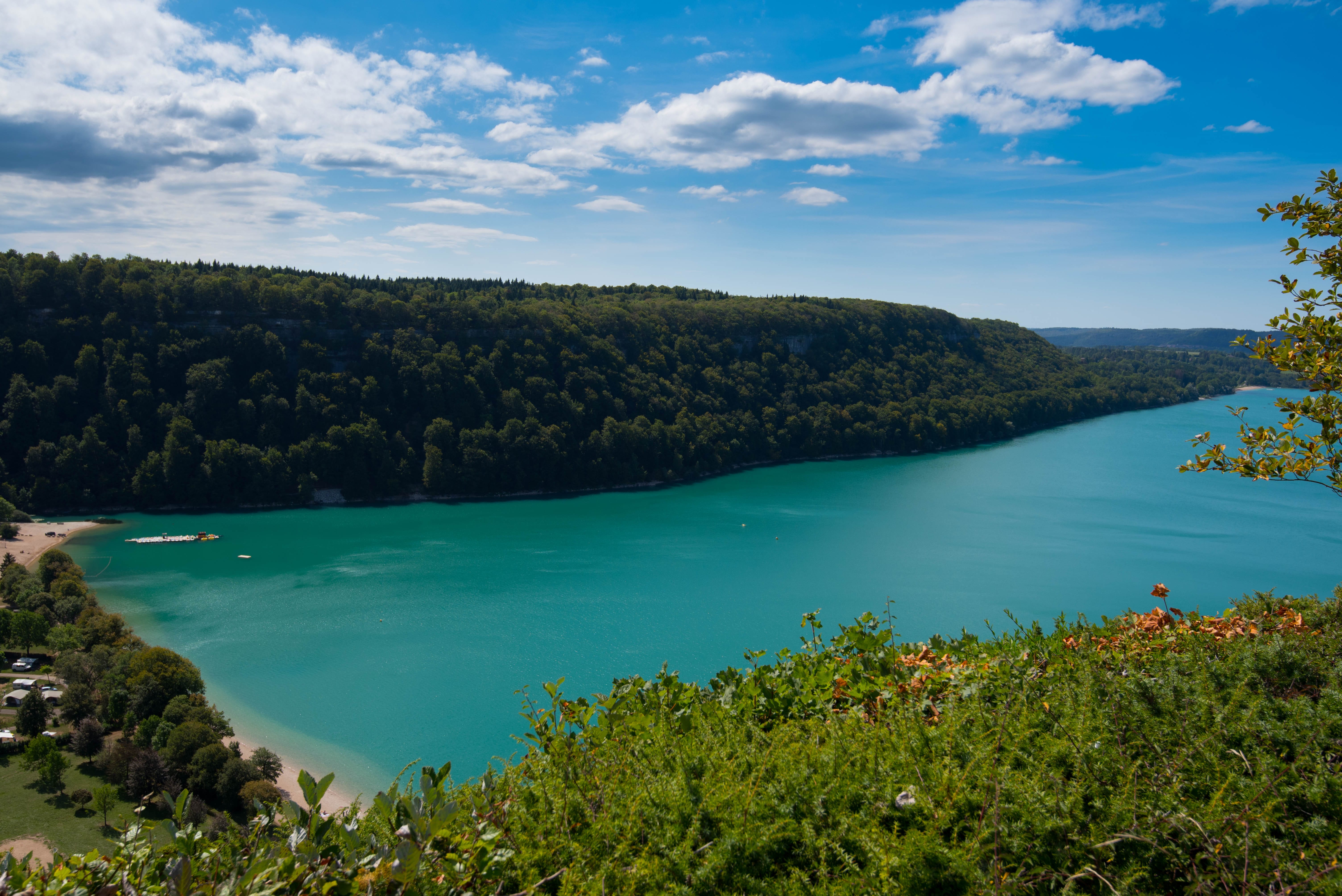 Een turkooisblauw meer, omringd door dichtbeboste heuvels, met een zonnige hemel en verspreide wolken erboven.