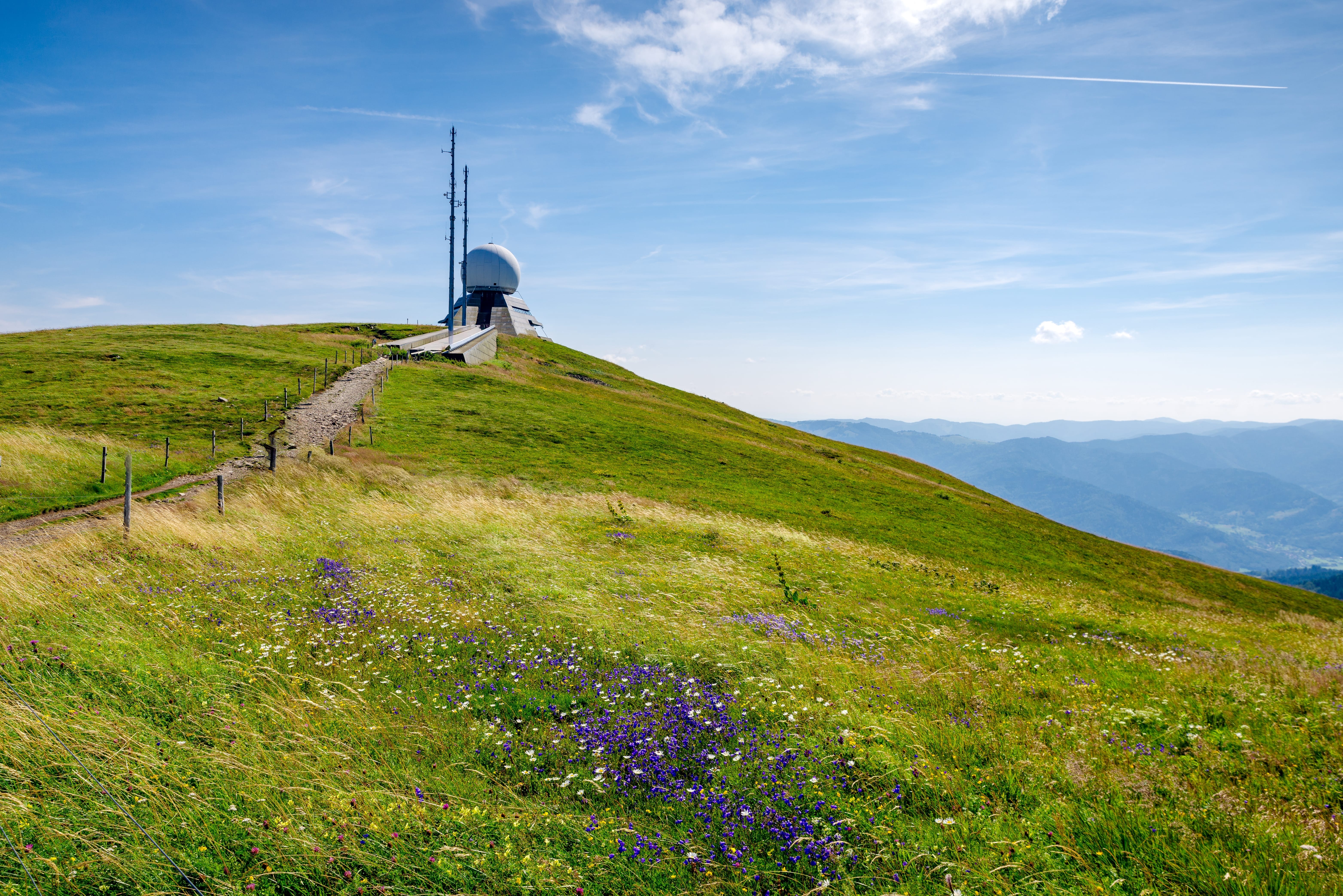 Een radarstation boven op een met gras en verspreide wilde bloemen begroeide heuvel; op de achtergrond bergen en een helderblauwe lucht met condenssporen.