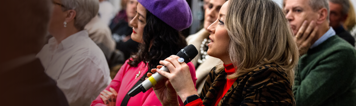 Woman in a red shirt and tiger-print jacket holding a microphone, seated among people at an event, listening attentively.