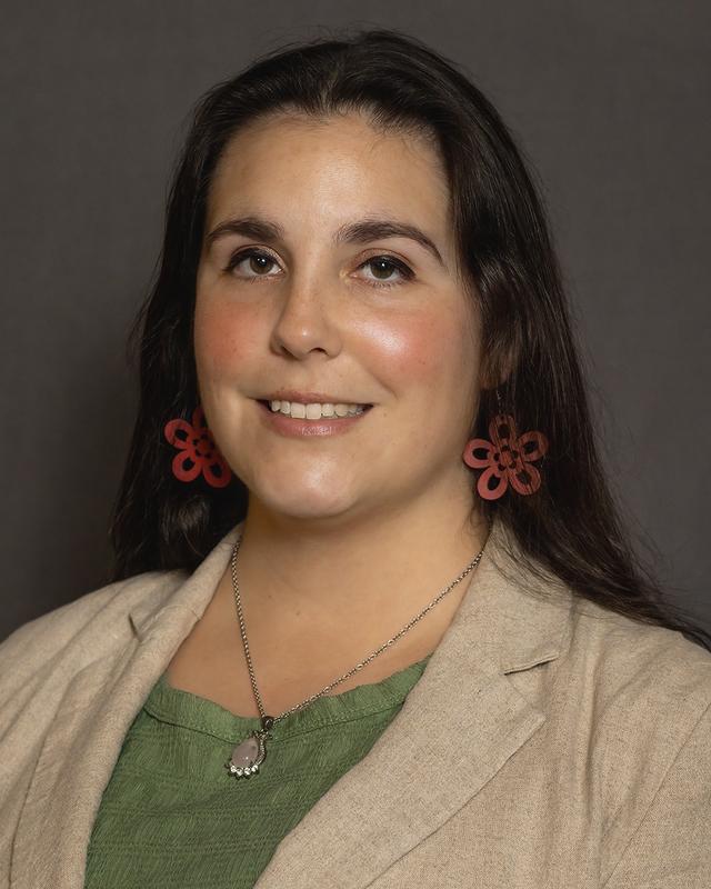 A woman with long dark hair, wearing red floral earrings, a green top, beige blazer, and a necklace, smiles against a neutral background.