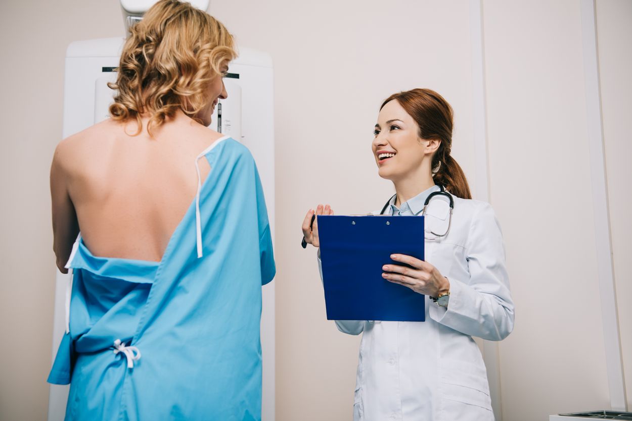 Eine Frau befindet sich bei der Mammographie und steht vor dem Röntgengerät. Sie trägt einen Patientenkittel in Blau und die behandelnde Ärztin steht neben ihr.