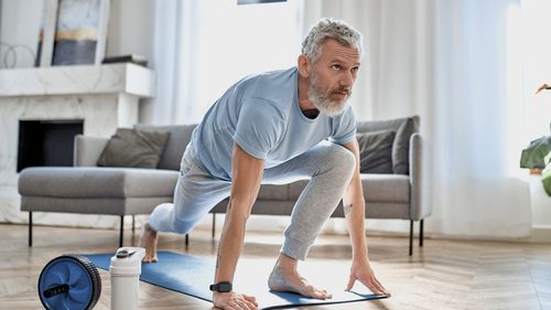 Ein Mann mit grauen Haaren mach eine Yogaübung in seinem Wohnzimmer auf einer Yogamatte.