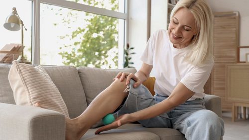 Eine blonde Frau sitzt auf dem Sofa und rollt mit einem Ball über ihre Wade.