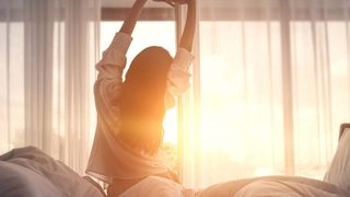 Frau streckt sich morgens entspannt im Bett, weiches Sonnenlicht fällt durch die Vorhänge und vermittelt ein Gefühl von erholsamem Schlaf.