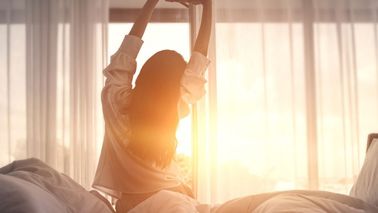 Frau streckt sich morgens entspannt im Bett, weiches Sonnenlicht fällt durch die Vorhänge und vermittelt ein Gefühl von erholsamem Schlaf.