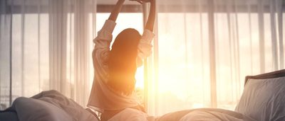 Frau streckt sich morgens entspannt im Bett, weiches Sonnenlicht fällt durch die Vorhänge und vermittelt ein Gefühl von erholsamem Schlaf.