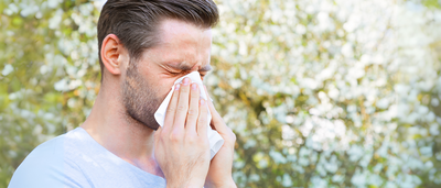 Mann im T-Shirt schnäuzt sich im Freien mit einem Taschentuch – zeigt typische Symptome eines grippalen Infekts im Sommer