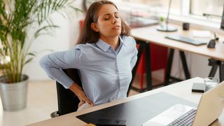 Frau mit Rückenschmerzen sitzt am Schreibtisch im Büro und hält sich den unteren Rücken nach langem Sitzen am Laptop.