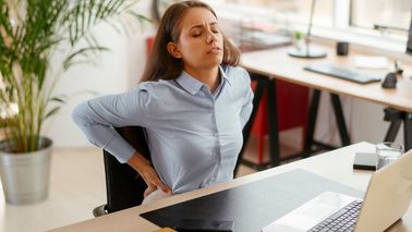 Frau mit Rückenschmerzen sitzt am Schreibtisch im Büro und hält sich den unteren Rücken nach langem Sitzen am Laptop.