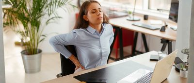 Frau mit Rückenschmerzen sitzt am Schreibtisch im Büro und hält sich den unteren Rücken nach langem Sitzen am Laptop.