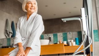 Ältere Frau sitzt lächelnd am Badewannenrand – sie wirkt sicher in ihrem modernen Badezimmer.
