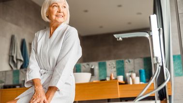 Ältere Frau sitzt lächelnd am Badewannenrand – sie wirkt sicher in ihrem modernen Badezimmer.