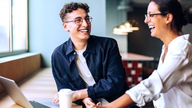 Zwei Kollegen lachen gemeinsam bei der Arbeit am Schreibtisch – entspannte Atmosphäre trotz Alltagsstress