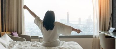 Frau sitzt morgens im Hotelbett und streckt sich nach einer Nacht im Urlaub