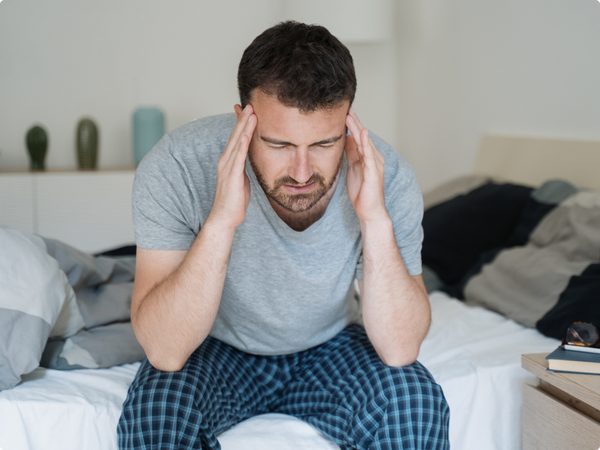 Mann sitzt am Bett und fasst sich an die Schläfe