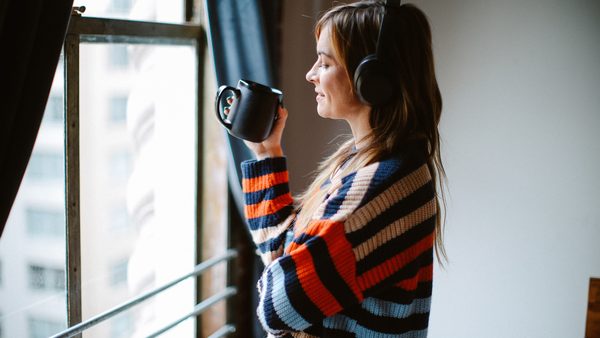 Frau steht entspannt mit einer Tasse Tee am Fenster und hört Musik