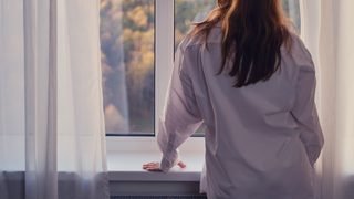 Frau steht am Fenster und schaut auf einen Herbstwald.