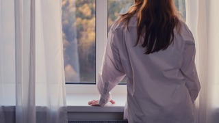 Frau steht am Fenster und schaut auf einen Herbstwald.