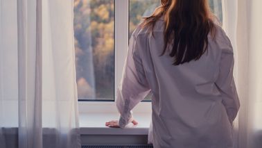 Frau steht am Fenster und schaut auf einen Herbstwald.