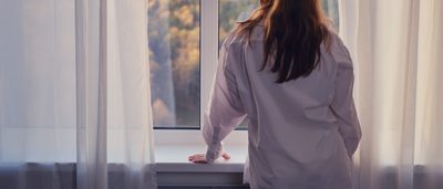 Frau steht am Fenster und schaut auf einen Herbstwald.