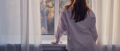 Frau steht am Fenster und schaut auf einen Herbstwald.