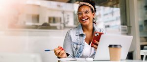 Lachende Frau mit Smartphone und Laptop im Café. Sie wirkt energiegeladen – Tipps gegen Müdigkeit für mehr Schwung im Alltag.