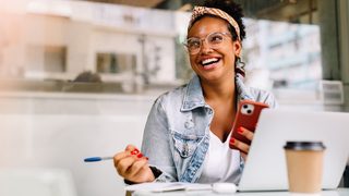 Lachende Frau mit Smartphone und Laptop im Café. Sie wirkt energiegeladen – Tipps gegen Müdigkeit für mehr Schwung im Alltag.