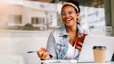 Lachende Frau mit Smartphone und Laptop im Café. Sie wirkt energiegeladen – Tipps gegen Müdigkeit für mehr Schwung im Alltag.