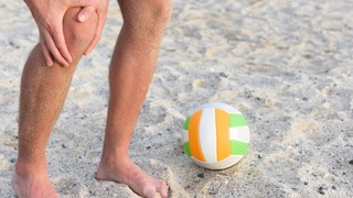 Mann mit Springerknie steht im Sand und fasst sich ans Knie.