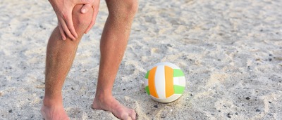 Mann mit Springerknie steht im Sand und fasst sich ans Knie.