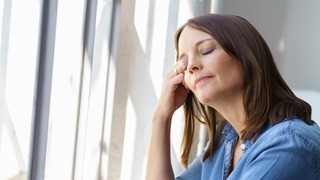Frau steht mit geschlossenen Augen an einem Fenster.