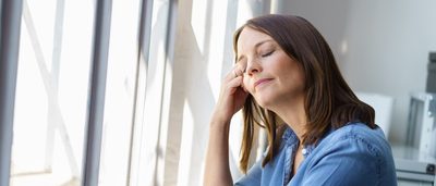 Frau steht mit geschlossenen Augen an einem Fenster.