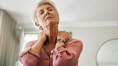 Ältere Frau auf einer Couch fasst sich an den Hals.