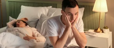 Mann sitzt auf einem Bett und hat Schlafstörungen durch Stress.