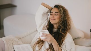 Frau entspannt auf dem Sofa mit Buch und Tee – Stressbewältigung zur Unterstützung des weiblichen Zyklus.