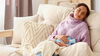 Entspannte Frau mit Wärmflasche auf dem Sofa – Wärme kann bei einer Blasenentzündung helfen.